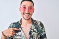 Young handsome man wearing Hawaiian sumer shirt and pink sunglasses over isolated background with surprise face pointing finger to Royalty Free Stock Photo