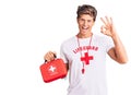 Young handsome man wearing lifeguard t shirt holding first aid kit doing ok sign with fingers, smiling friendly gesturing