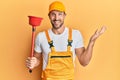 Young handsome man wearing plumber uniform holding toilet plunger celebrating achievement with happy smile and winner expression Royalty Free Stock Photo