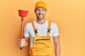 Young handsome man wearing plumber uniform holding toilet plunger looking positive and happy standing and smiling with a confident Royalty Free Stock Photo