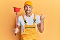 Young handsome man wearing plumber uniform holding toilet plunger screaming proud, celebrating victory and success very excited