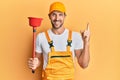 Young handsome man wearing plumber uniform holding toilet plunger smiling happy pointing with hand and finger to the side Royalty Free Stock Photo