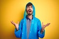 Young handsome man wearing rain coat with hood standing over isolated yellow background crazy and mad shouting and yelling with