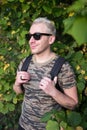 A young handsome man wearing t-shirt and black sunglasses on background of green and yellow leaves. Royalty Free Stock Photo