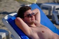 A young handsome man of 25-30 years old, a brunette, in weed sunglasses, lies relaxed on a lounger on a sandy sea beach, takes a Royalty Free Stock Photo