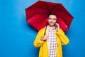 Young handsome bearded man in yellow raincoat with red umbrella talking by mobile phone  over blue background Royalty Free Stock Photo