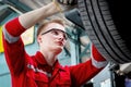 Young handsome mechanic in red uniform working with lifted vehicle, auto mechanic checking wheel at garage, technician check, Royalty Free Stock Photo