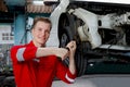 Young handsome mechanic in red uniform working with lifted vehicle, auto mechanic checking wheel at garage, technician check, Royalty Free Stock Photo