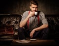 Young handsome old-fashioned bearded man with cup of coffee sitting on comfortable leather sofa on dark background. Royalty Free Stock Photo