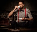 Young handsome old-fashioned bearded man with cup of coffee sitting on comfortable leather sofa on dark background. Royalty Free Stock Photo