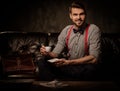 Young handsome old-fashioned bearded man with cup of coffee sitting on comfortable leather sofa on dark background.