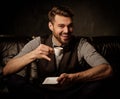 Young handsome old-fashioned bearded man having fun with cup of coffee on comfortable leather sofa on dark background. Royalty Free Stock Photo