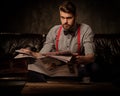 Young handsome old-fashioned bearded man with newspaper sitting on comfortable leather sofa on dark background. Royalty Free Stock Photo