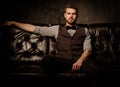 Young handsome old-fashioned bearded man sitting on comfortable leather sofa on dark background. Royalty Free Stock Photo