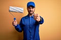 Young handsome painter man with beard wearing blue uniform and cap painting using roller annoyed and frustrated shouting with Royalty Free Stock Photo