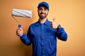 Young handsome painter man with beard wearing blue uniform and cap painting using roller happy with big smile doing ok sign, thumb Royalty Free Stock Photo