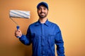 Young handsome painter man with beard wearing blue uniform and cap painting using roller with a happy face standing and smiling Royalty Free Stock Photo