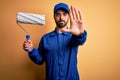 Young handsome painter man with beard wearing blue uniform and cap painting using roller with open hand doing stop sign with Royalty Free Stock Photo