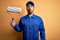 Young handsome painter man with beard wearing blue uniform and cap painting using roller scared in shock with a surprise face, Royalty Free Stock Photo