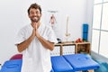 Young handsome physiotherapist man working at pain recovery clinic praying with hands together asking for forgiveness smiling Royalty Free Stock Photo