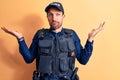 Young handsome policeman wearing police uniform and bulletprof over yellow background clueless and confused with open arms, no Royalty Free Stock Photo