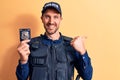 Young handsome policeman wearing uniform and bulletprof holding police badge pointing thumb up to the side smiling happy with open