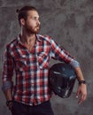 Young handsome redhead biker in a flannel shirt, holds motorcycle helmet, posing on a gray background.