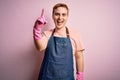 Young handsome redhead cleaner man doing housework wearing apron and gloves pointing finger up with successful idea Royalty Free Stock Photo