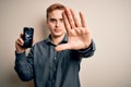 Young handsome redhead man holding broken smartphone showing cracked screen with open hand doing stop sign with serious and Royalty Free Stock Photo