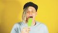 Young handsome satisfied man with dreadlocks in black cap eating ice cream on yellow background. Joyful guy in blue t Royalty Free Stock Photo