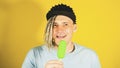 Young handsome satisfied man with dreadlocks in black cap eating ice cream on yellow background. Joyful guy in blue t Royalty Free Stock Photo