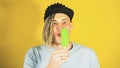 Young handsome satisfied man with dreadlocks in black cap eating ice cream on yellow background. Joyful guy in blue t Royalty Free Stock Photo