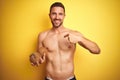 Young handsome shirtless man holding exotic tropical coconut over isolated yellow background with surprise face pointing finger to Royalty Free Stock Photo