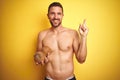 Young handsome shirtless man holding exotic tropical coconut over isolated yellow background very happy pointing with hand and Royalty Free Stock Photo