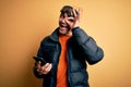 Young handsome skier man wearing ski goggles using smartphone over yellow background with happy face smiling doing ok sign with