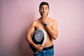 Young handsome slim man with beard shirtless holding weight machine over pink background cover mouth with hand shocked with shame