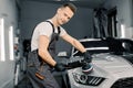 Young handsome smiling Caucasian man, worker of auto detailing service, holds a polisher in the hand and polishes the