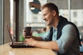 Young handsome smiling man working remotely in his laptop holdscup of hot coffee in cafe Royalty Free Stock Photo