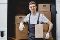 A young handsome smiling worker wearing uniform is standing next to the van full of boxes holding a box in his hands Royalty Free Stock Photo