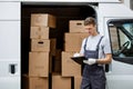 A young handsome smiling worker wearing uniform is standing next to the van full of boxes holding a clipboard in his Royalty Free Stock Photo