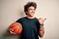 Young handsome sportsman holding basketball ball standing over isolated white background pointing and showing with thumb up to the Royalty Free Stock Photo
