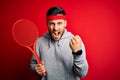 Young handsome sportsman holding tennis racket wearing sportswear over red background annoyed and frustrated shouting with anger,