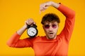 Young handsome surprised man points at alarm clock in his hand, isolated yellow background