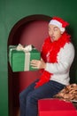 Young handsome surprised man wearing Santa hat showing on Christmas gift box in the green arch background