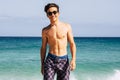 Young handsome teenager in swimsuit walking at the beach with blue sky and ocean water in background enjoying summer outdoor Royalty Free Stock Photo