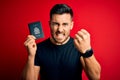 Young handsome tourist man holding canada canadian passport id over red background annoyed and frustrated shouting with anger,