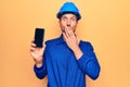 Young handsome worker man wearing uniform and hardhat holding smartphone showing screen covering mouth with hand, shocked and Royalty Free Stock Photo