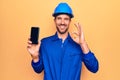 Young handsome worker man wearing uniform and hardhat holding smartphone showing screen doing ok sign with fingers, smiling Royalty Free Stock Photo