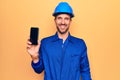 Young handsome worker man wearing uniform and hardhat holding smartphone showing screen looking positive and happy standing and Royalty Free Stock Photo