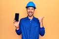 Young handsome worker man wearing uniform and hardhat holding smartphone showing screen pointing thumb up to the side smiling Royalty Free Stock Photo